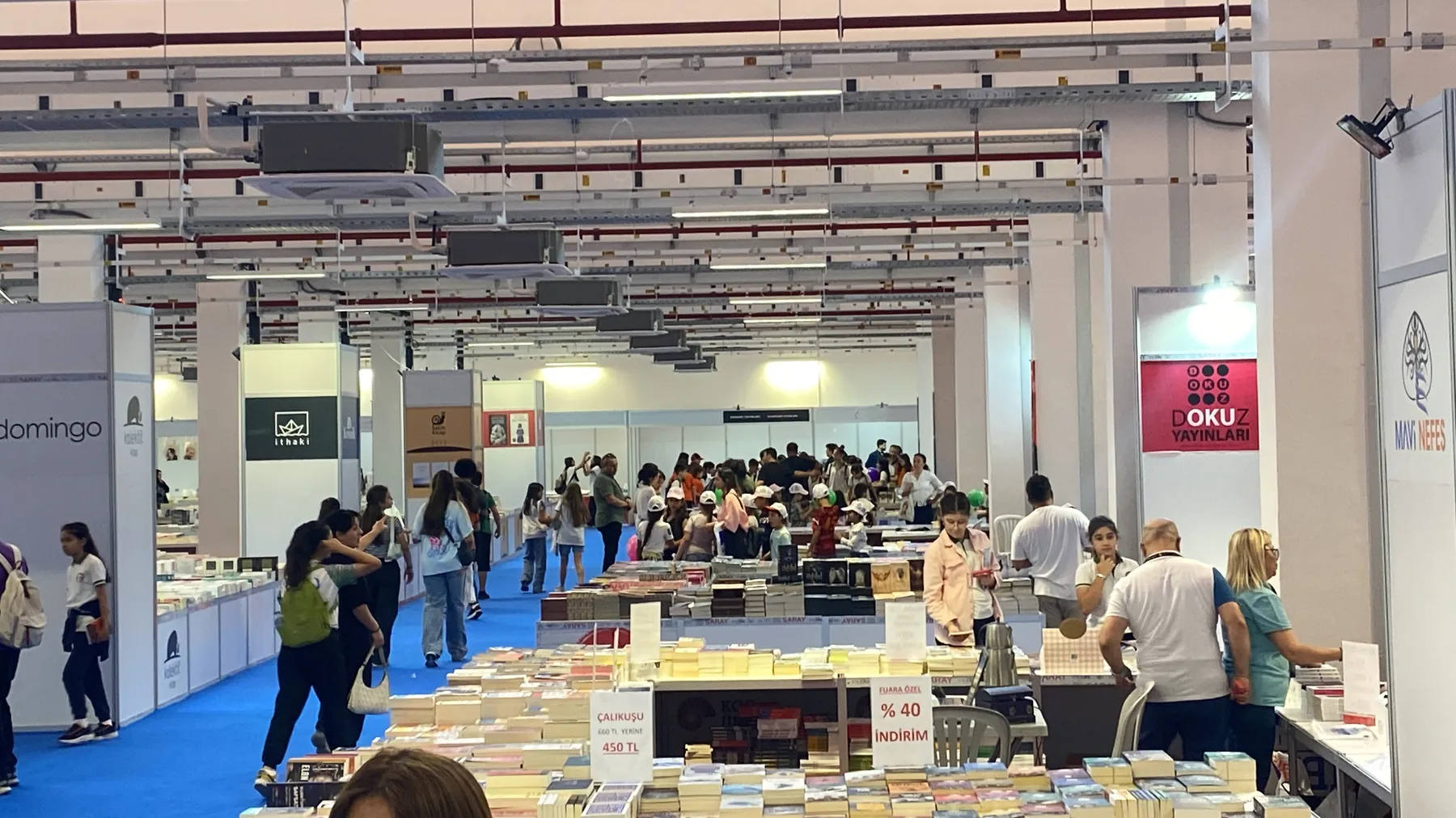 Fuar salonunun genel görünümü: mavi halı, sıralanmış stantlar ve kitap masaları